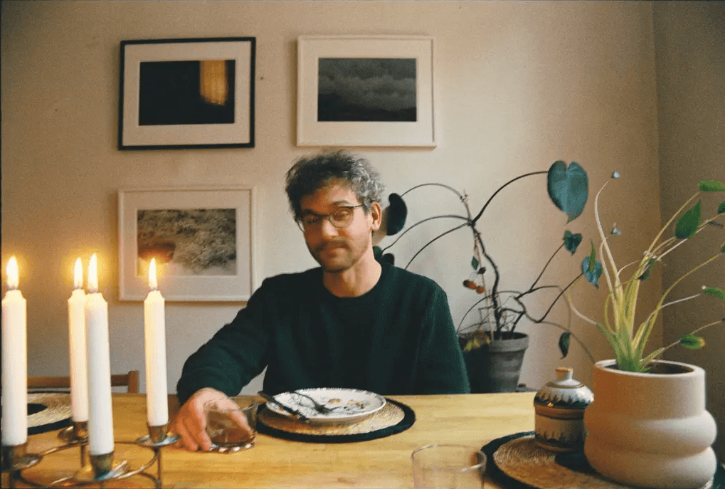 A person with glasses sits at a candlelit table with plants and framed pictures in the background.