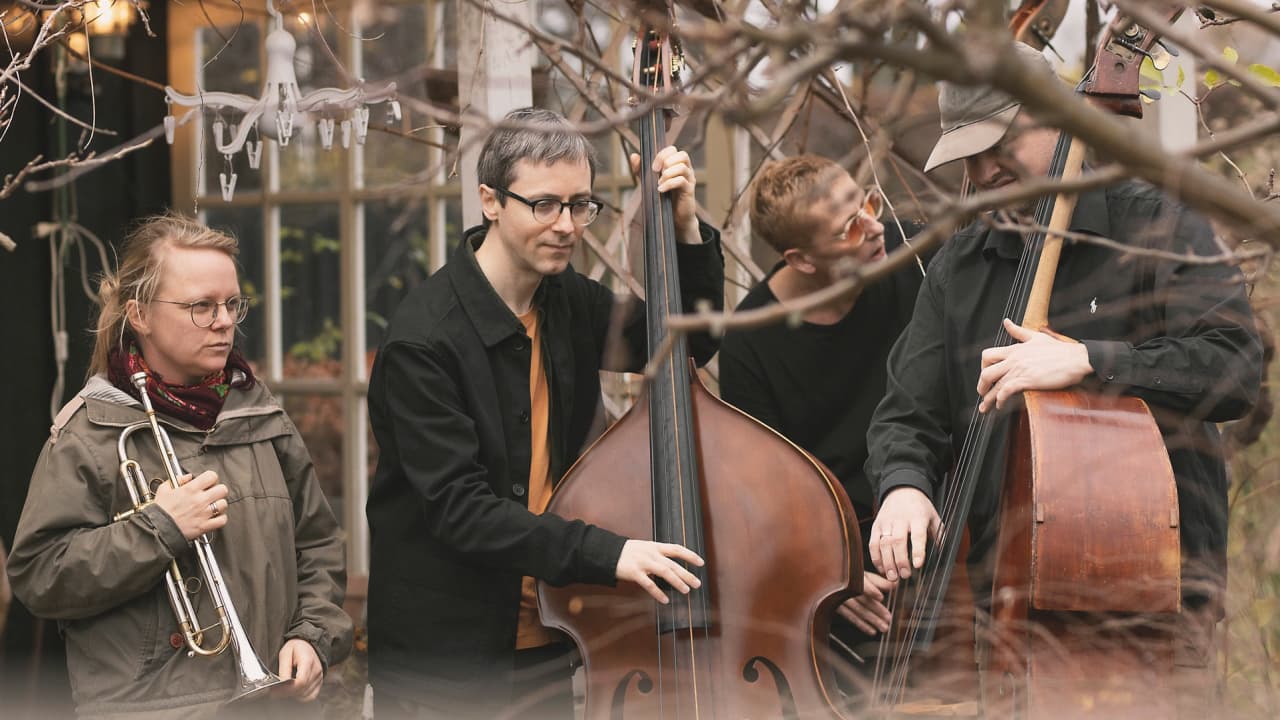 Four people are outdoors, one holding a trumpet and the others playing double basses, surrounded by branches.