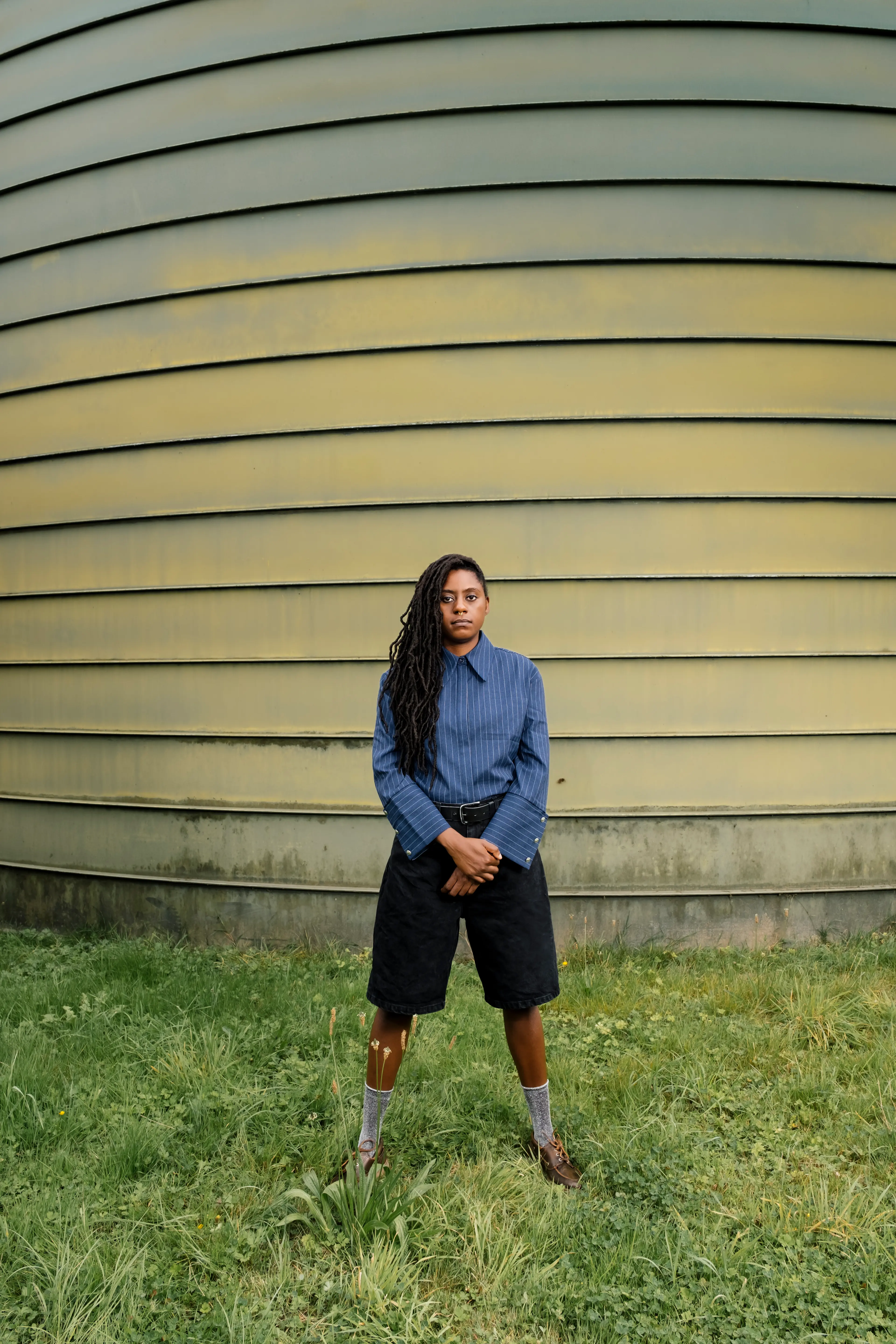 A person with long hair stands in front of a large, curved, yellow and green structure on a grassy area, wearing a blue shirt and black shorts.