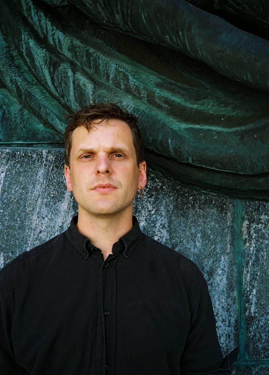 A person wearing a black shirt stands in front of a textured, greenish-blue background.