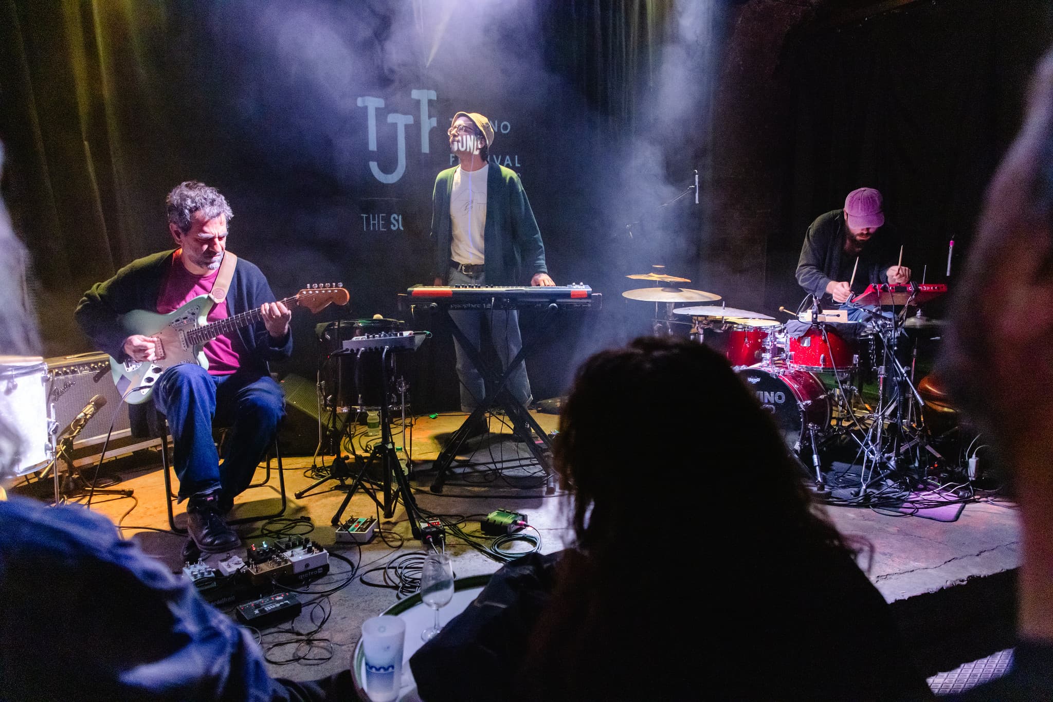 Three musicians are performing on stage with a guitarist, a keyboardist, and a drummer, surrounded by musical equipment, in front of an audience.