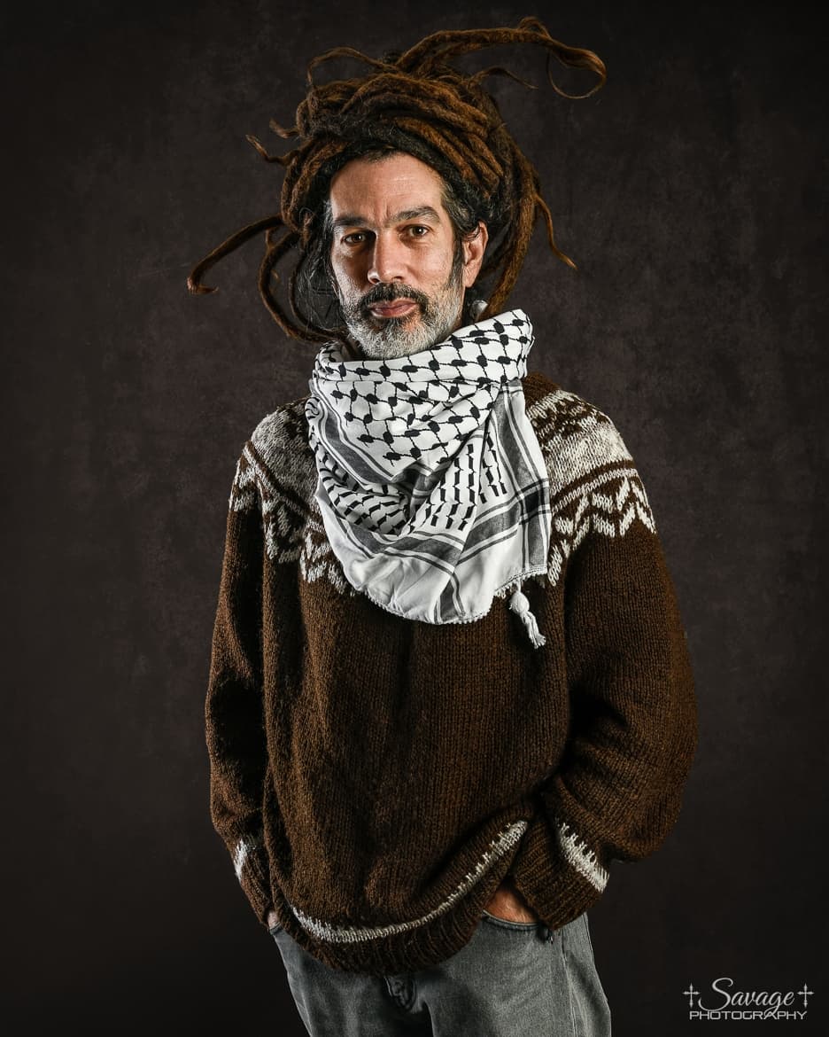 A person with dreadlocks is wearing a brown sweater and a patterned scarf, standing against a dark background.