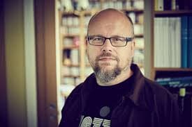 A man with glasses and a beard is standing indoors in front of shelves filled with books.