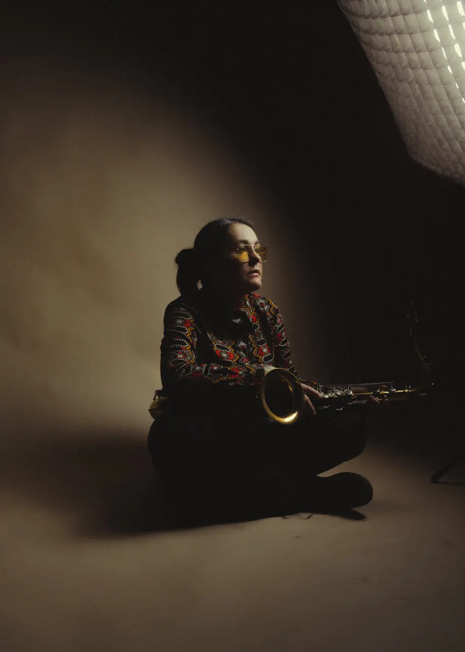 A person wearing sunglasses and a colorful outfit is sitting in a dimly lit space, holding a saxophone.