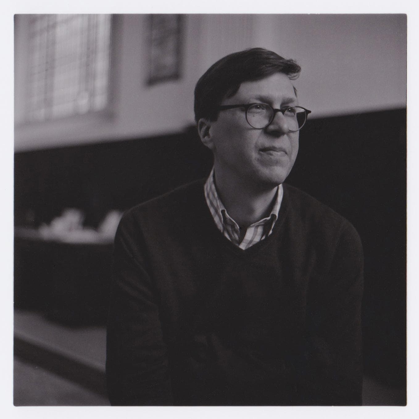 A man with glasses is sitting indoors, looking to the side, in a black and white photo.