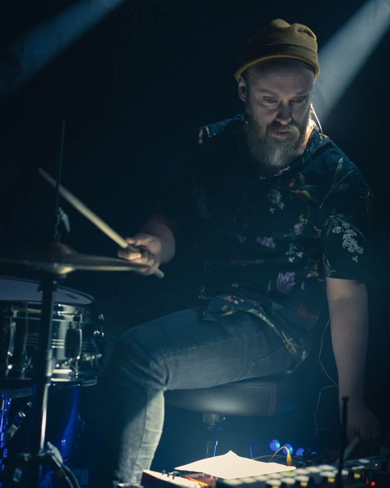 A person wearing a beanie is playing drums in dim lighting, illuminated by a spotlight.