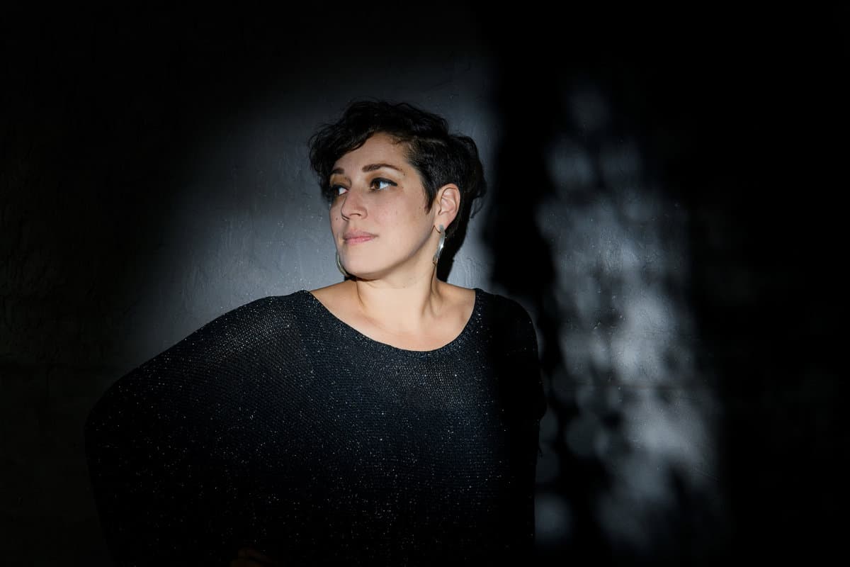 A person with short hair and hoop earrings is illuminated by a spotlight against a dark background.