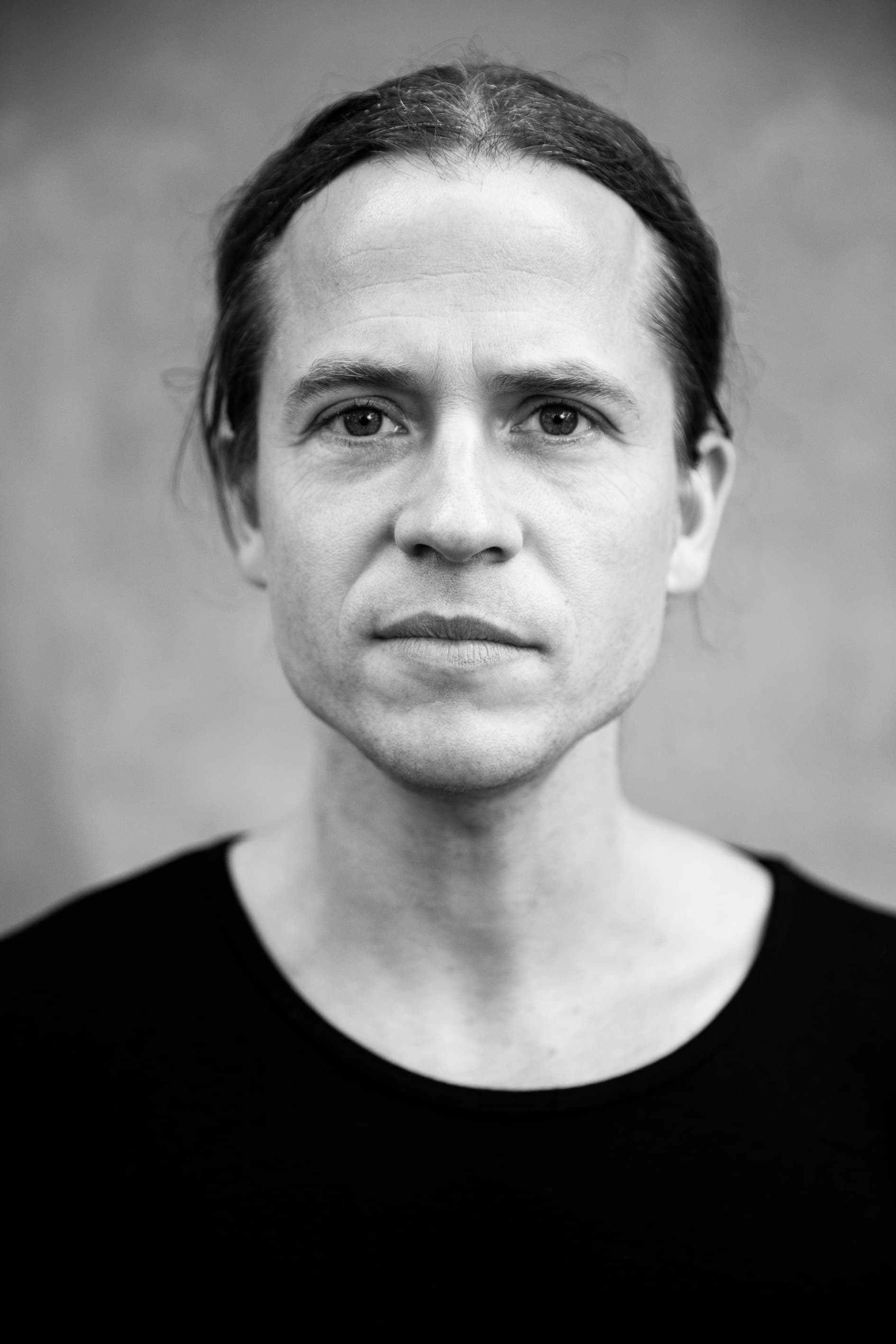 A black and white portrait of a person with long hair pulled back, wearing a black shirt, looking directly at the camera.