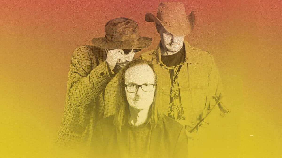 Three people in various hats and outfits stand against a gradient background, with the central figure wearing glasses.