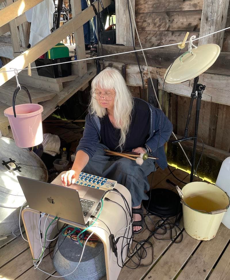 A person with long white hair is sitting by a laptop and audio equipment, surrounded by buckets and various tools, in a rustic wooden setting.
