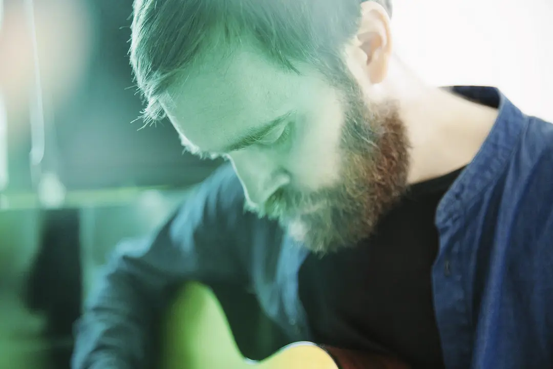 A bearded man is focused on playing a guitar, with a soft green light effect.