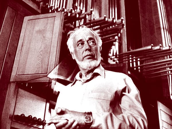 An older man with a beard stands in front of an organ, looking to the side.