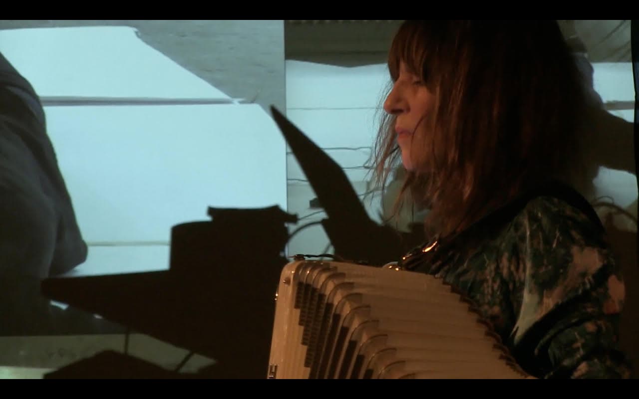 A person with long hair plays an accordion beside a projected image on a screen.