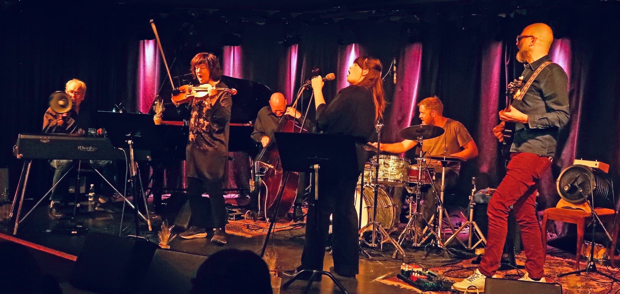 A band performs on stage with a keyboard, violin, double bass, singer, drums, and guitar.