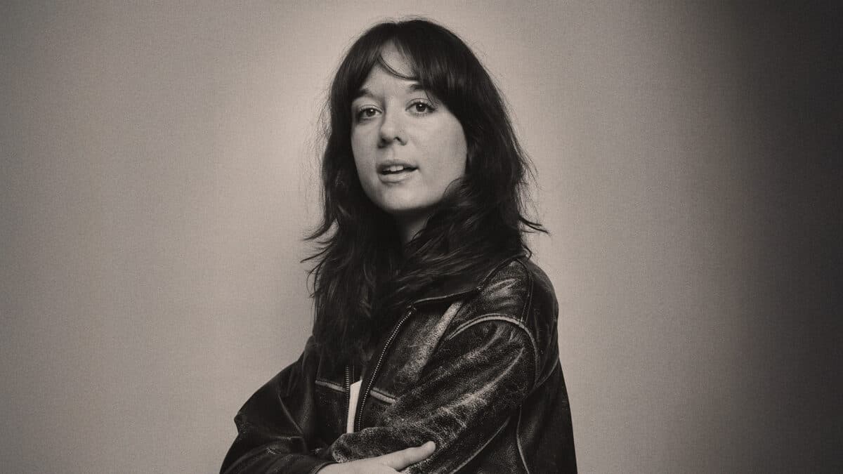 A person with long dark hair is wearing a leather jacket and looking at the camera against a neutral background.