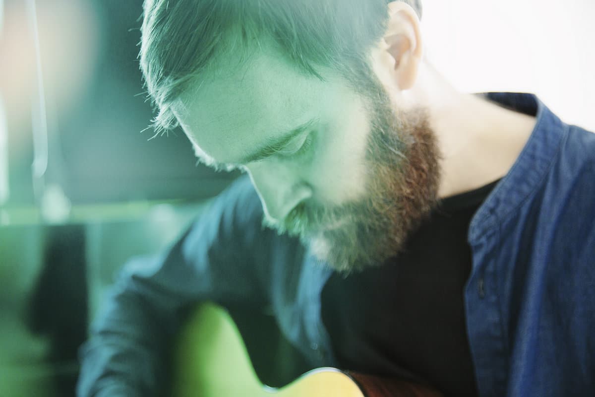 A person with a beard is playing a guitar, looking down with a soft greenish tint over the scene.