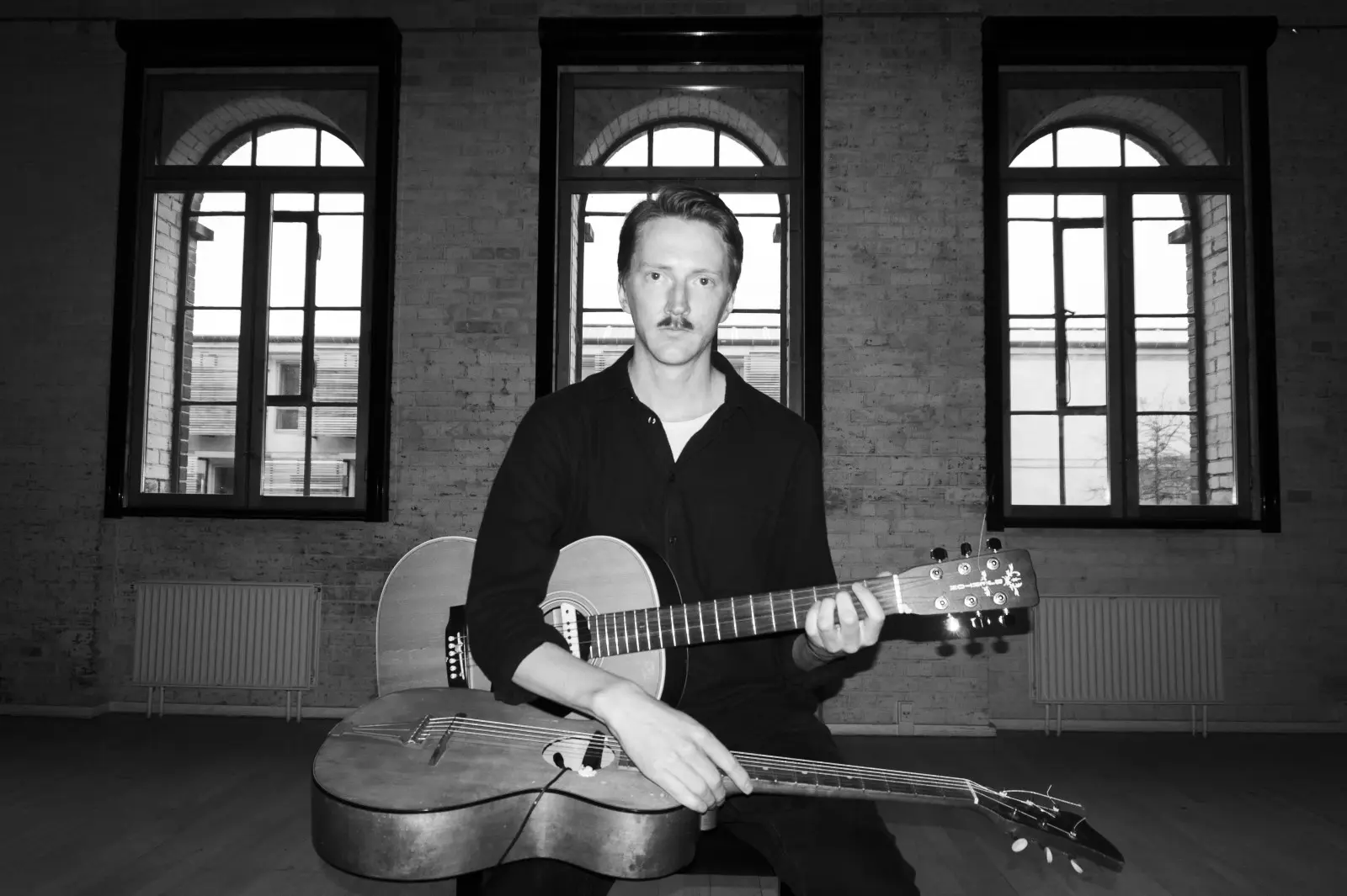 A person is sitting indoors, holding a guitar, with large windows in the background.