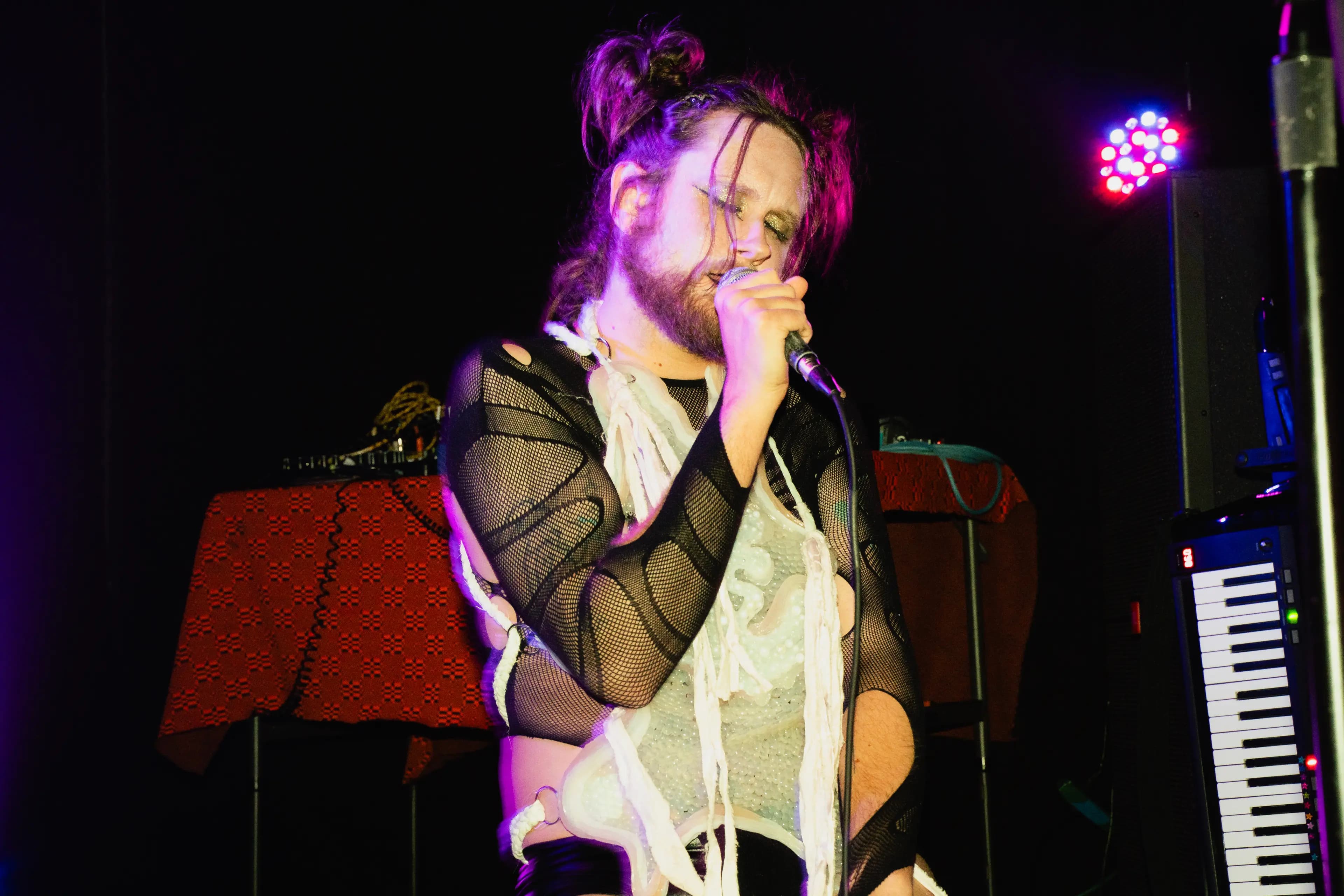 A person with colorful lighting sings into a microphone wearing a unique mesh and fabric outfit.