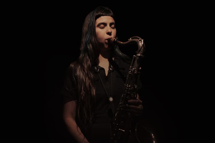 A person with long hair is playing a saxophone in a dimly lit setting.