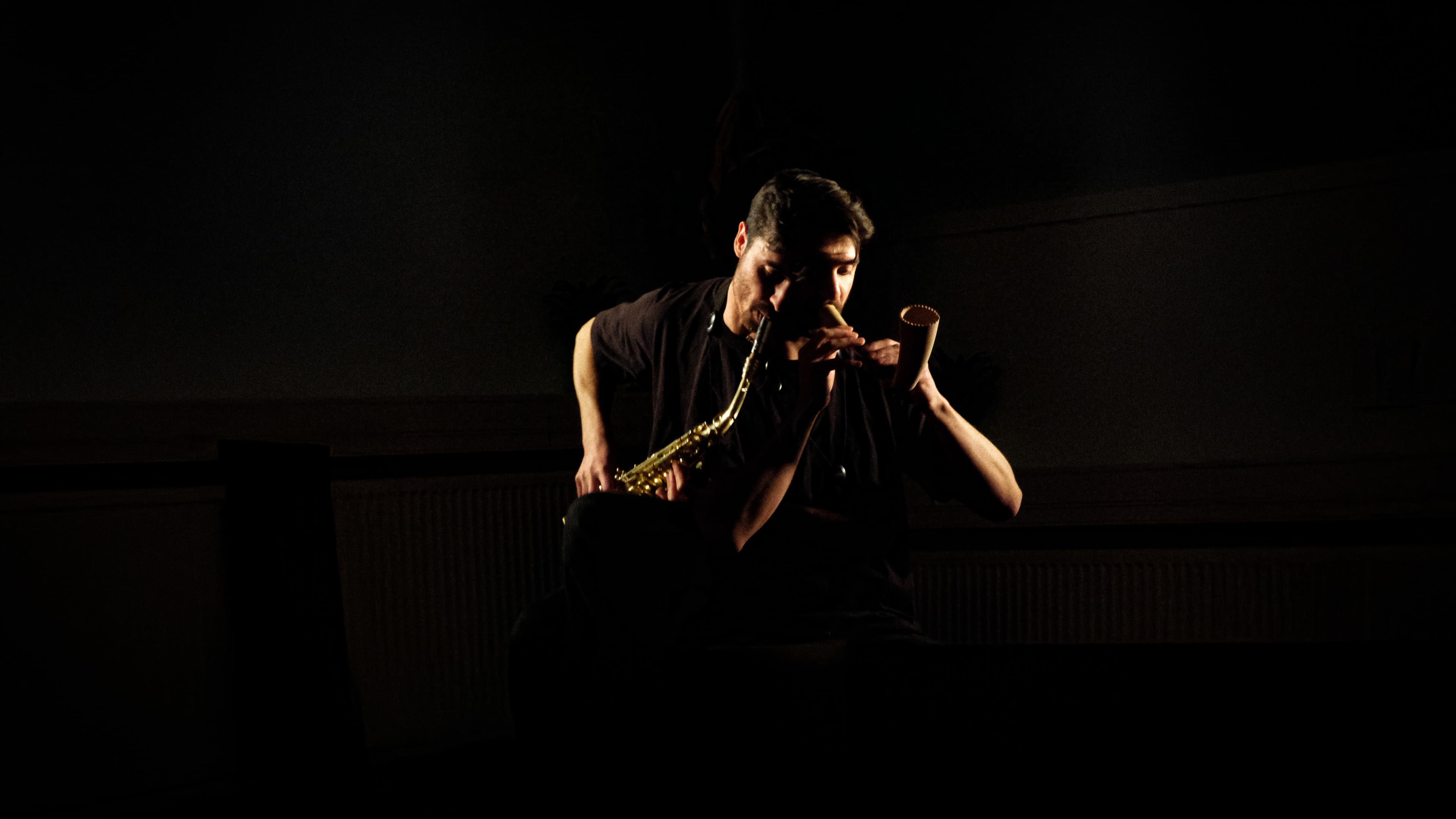 A person is playing a saxophone and a small wind instrument in a dimly lit setting.