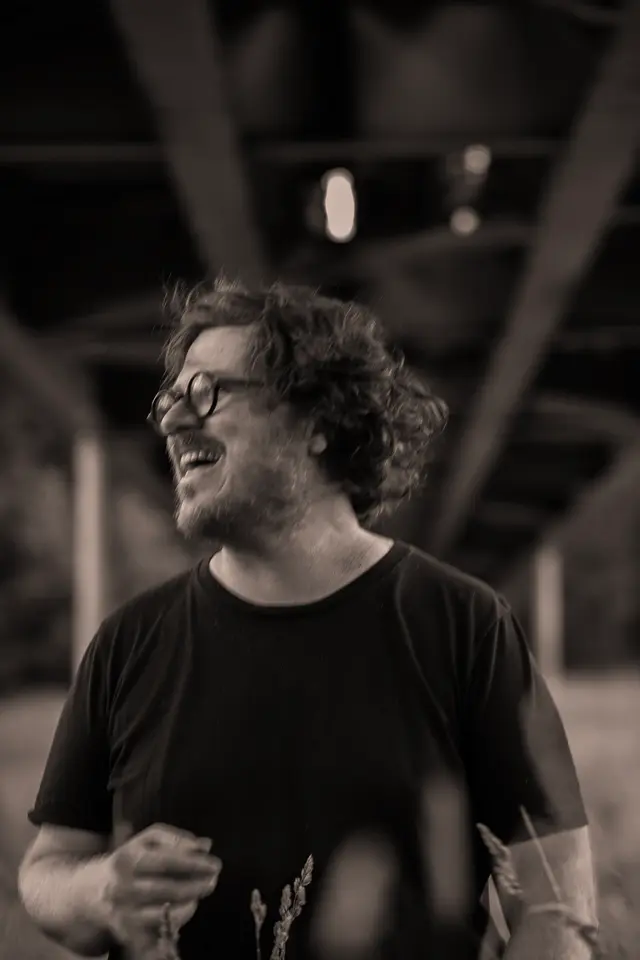 A person with curly hair and glasses is smiling broadly, standing outdoors under a bridge in a black and white photo.