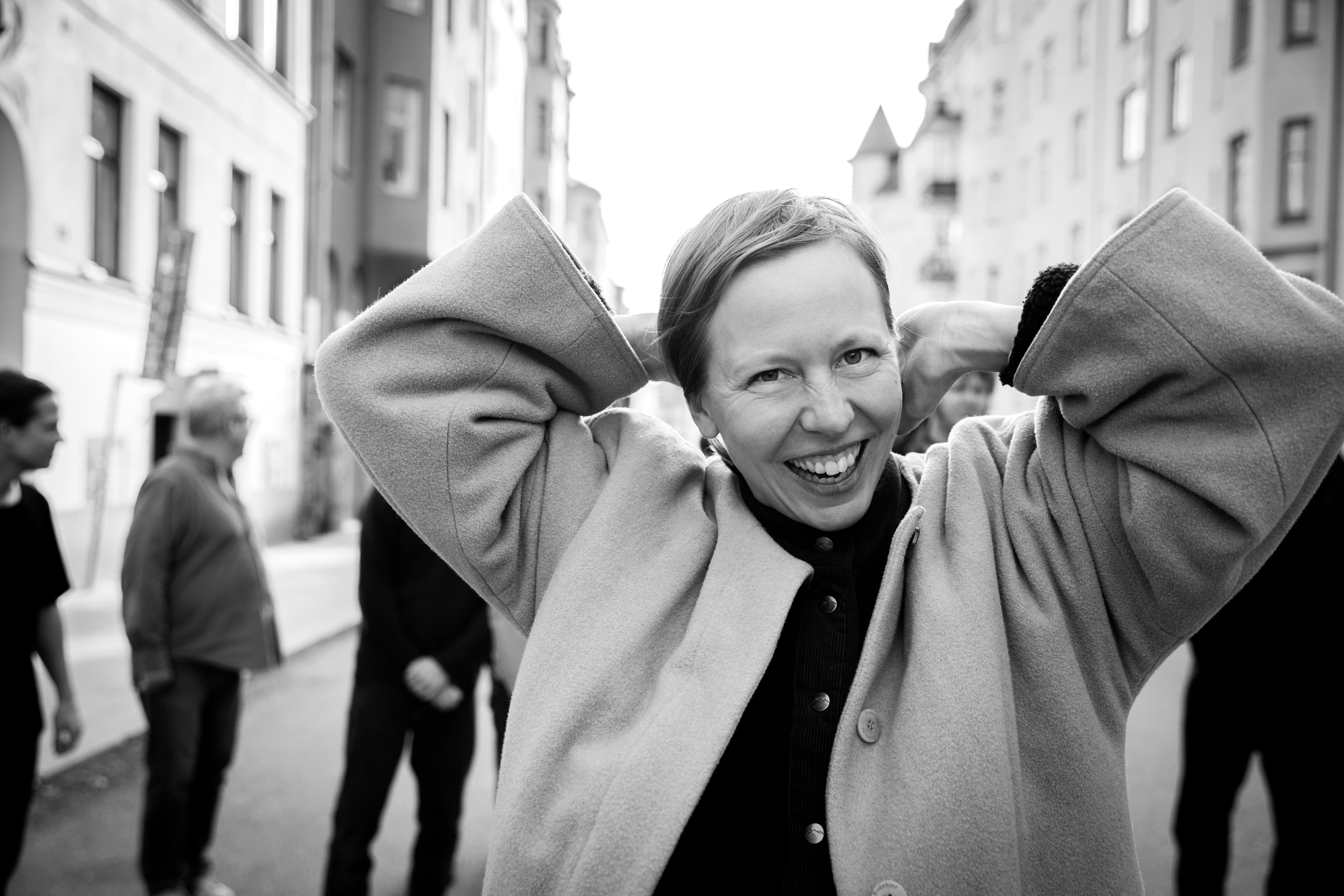 A person with short hair is smiling and adjusting their coat, with others blurred in the background on a city street.
