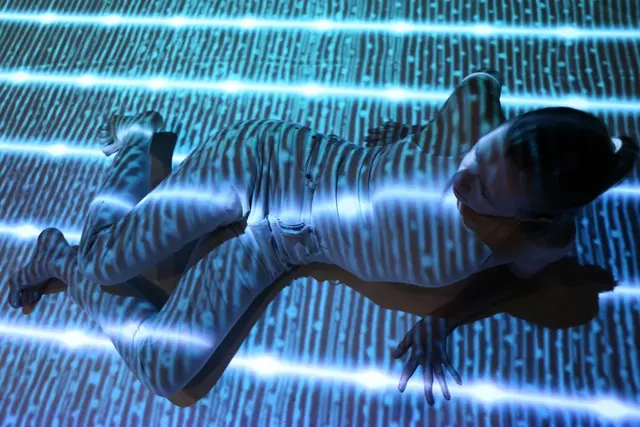 A person is lying on the floor with blue and white striped light projections covering their body and the ground.