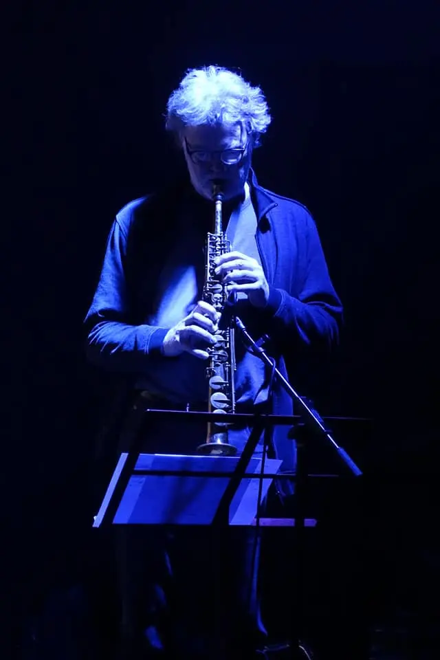 A person is playing a soprano saxophone under moody blue lighting with a music stand in front.